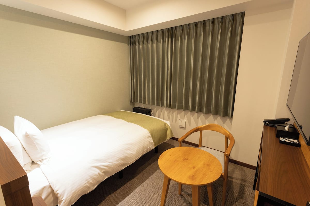 Una habitación de hotel con cama, una silla, escritorio, televisión y ventana con cortinas.