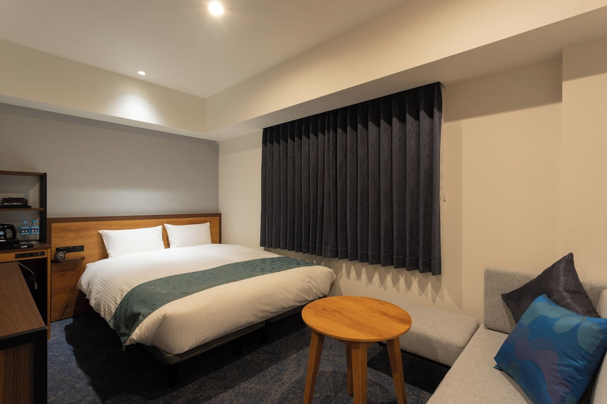 Habitación de hotel con cama, mesita de noche de madera, escritorio, mesa redonda de madera, sofá y ventana con cortinas.