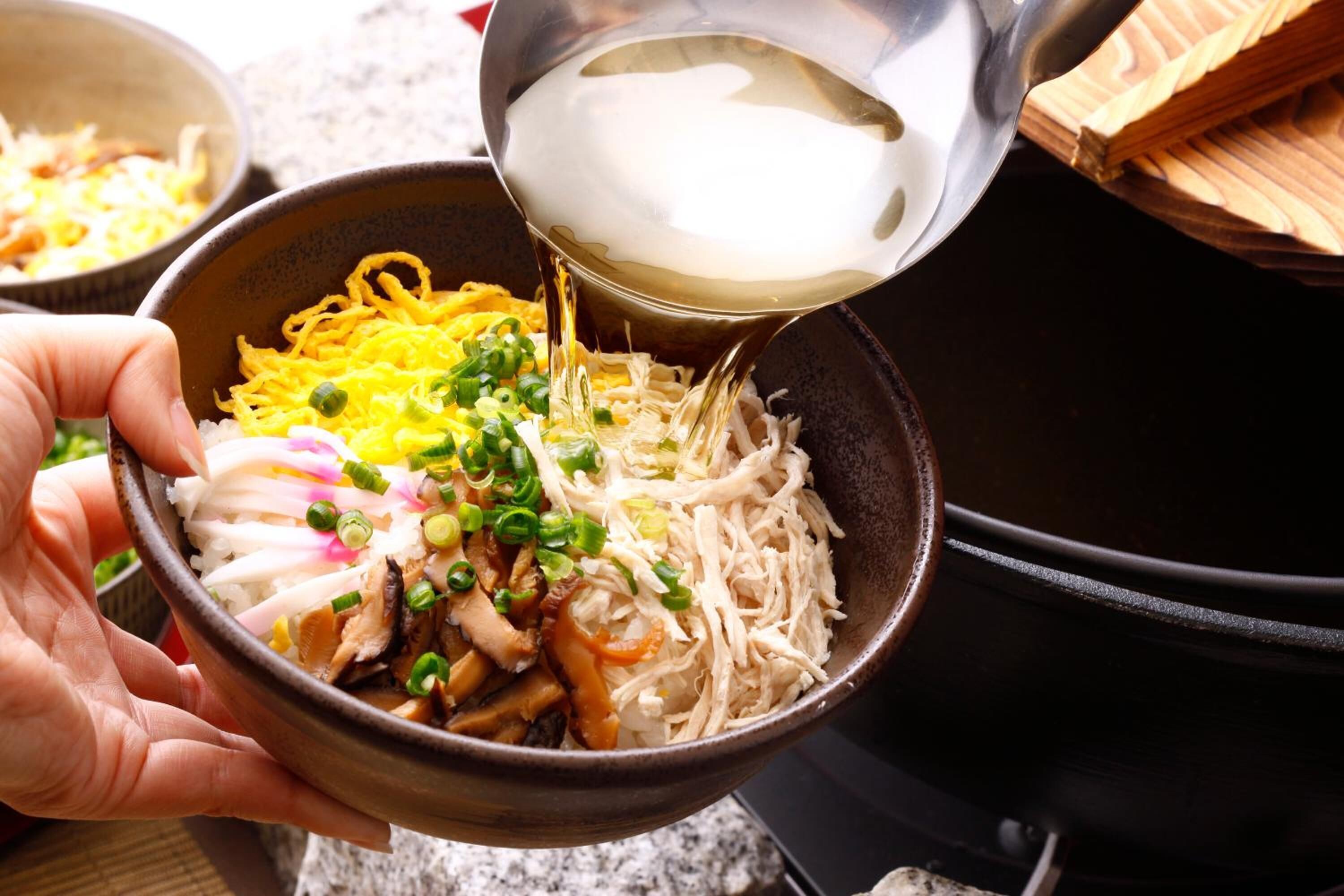 A bowl of noodles being topped with a clear liquid, likely broth, and garnished with green onions.