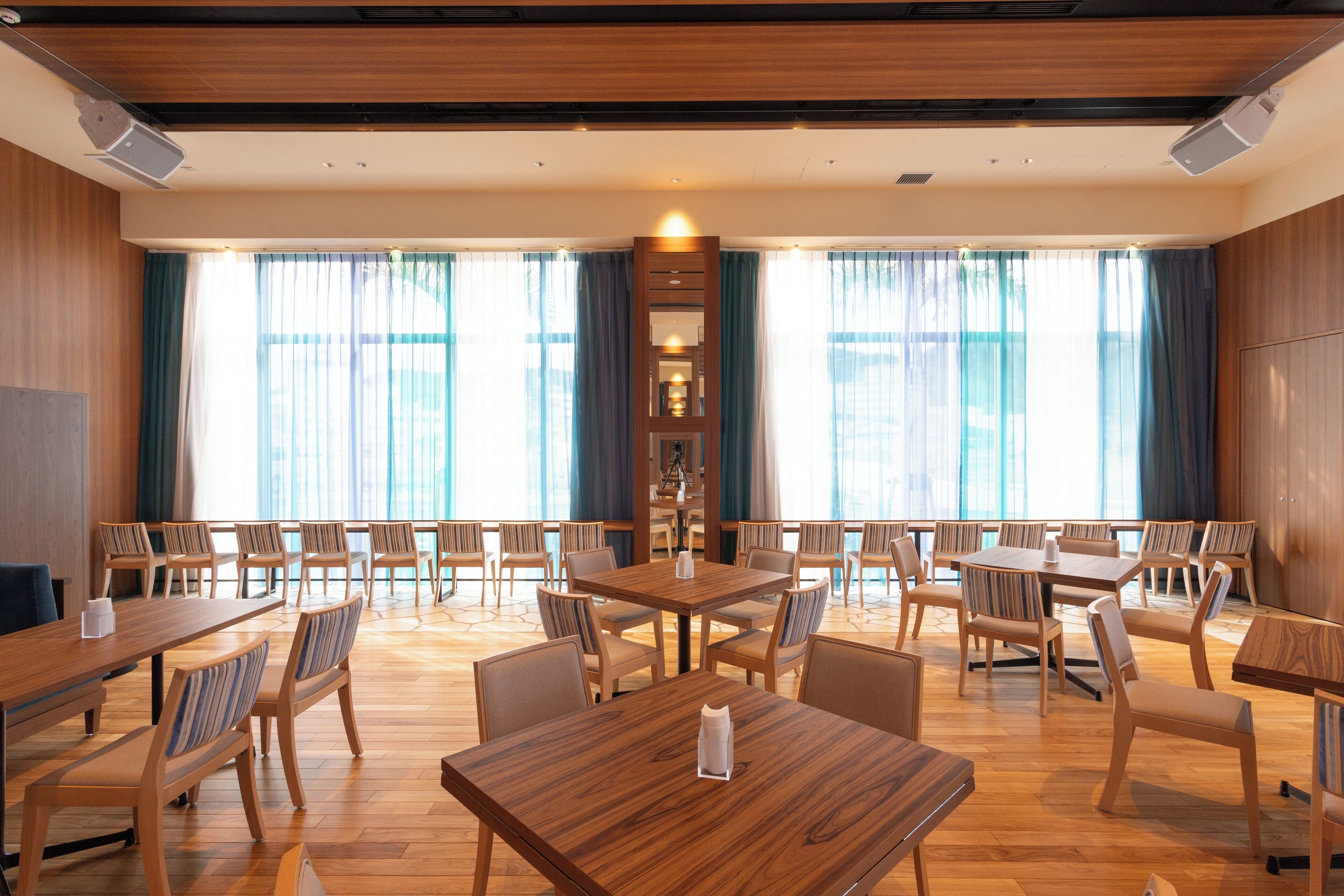 A spacious dining area with multiple wooden tables and chairs, large windows with curtains, and a central decorative column.
