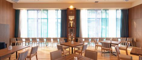 A spacious dining area with multiple wooden tables and chairs, large windows with curtains, and a central decorative column.