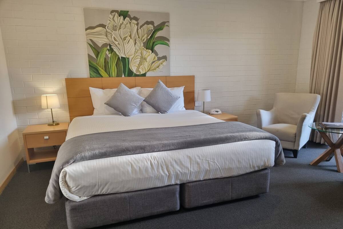 A hotel room with a large bed, a headboard, a nightstand with a lamp, a chair, and a glass table.