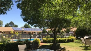 An outdoor patio with a glass table and chairs, a swimming pool, and solar panels on a building in the background. - Nagambie Motor Inn (Nagambie)