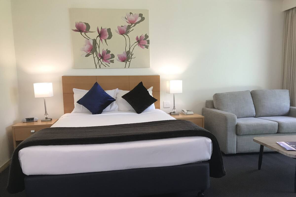 A hotel room with a bed, two bedside tables with lamps, a grey sofa, and a painting of flowers on the wall.