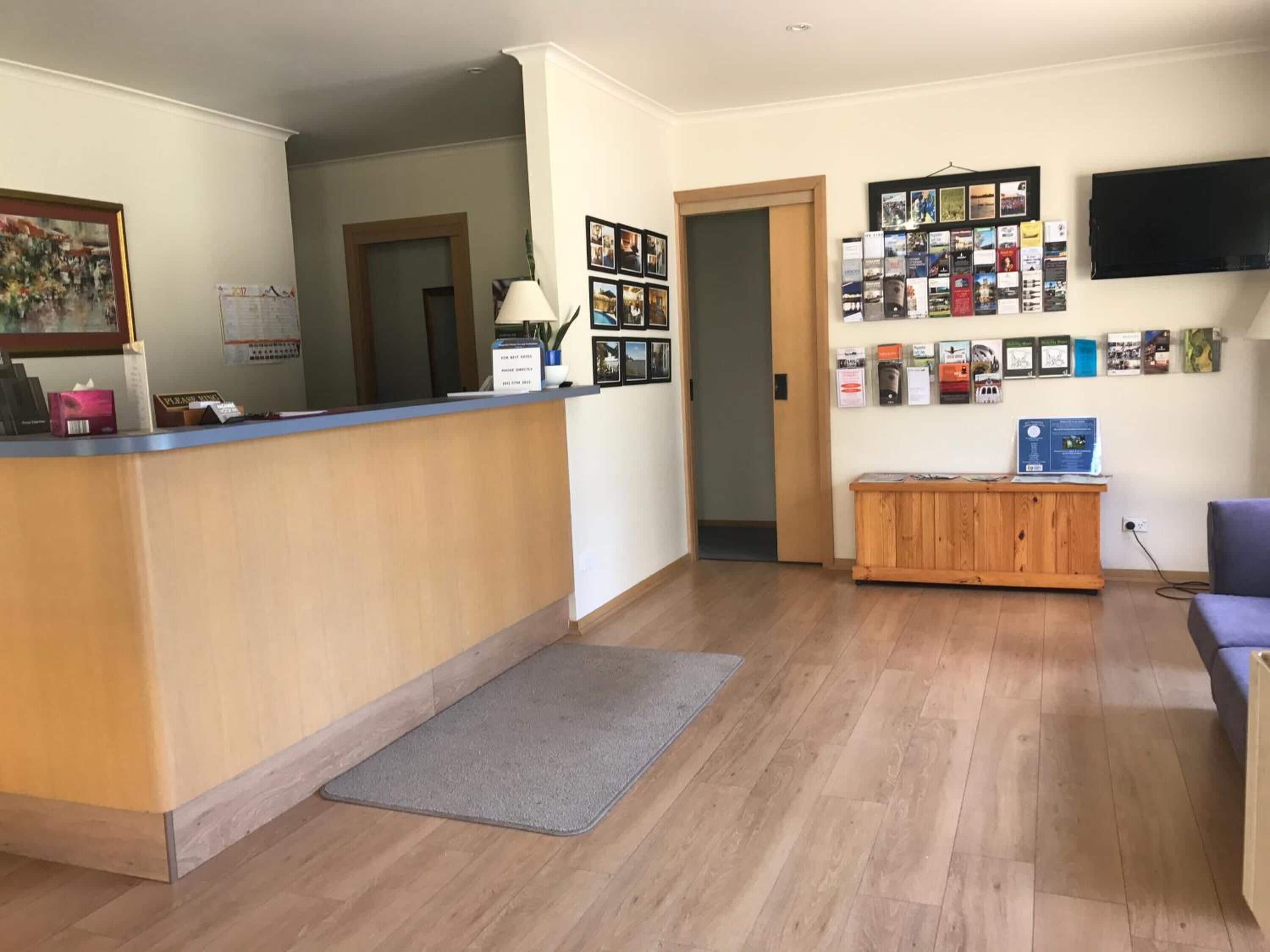 A reception area with a wooden reception desk, a grey mat, a wooden cabinet, and a wall with framed pictures and a mounted TV.