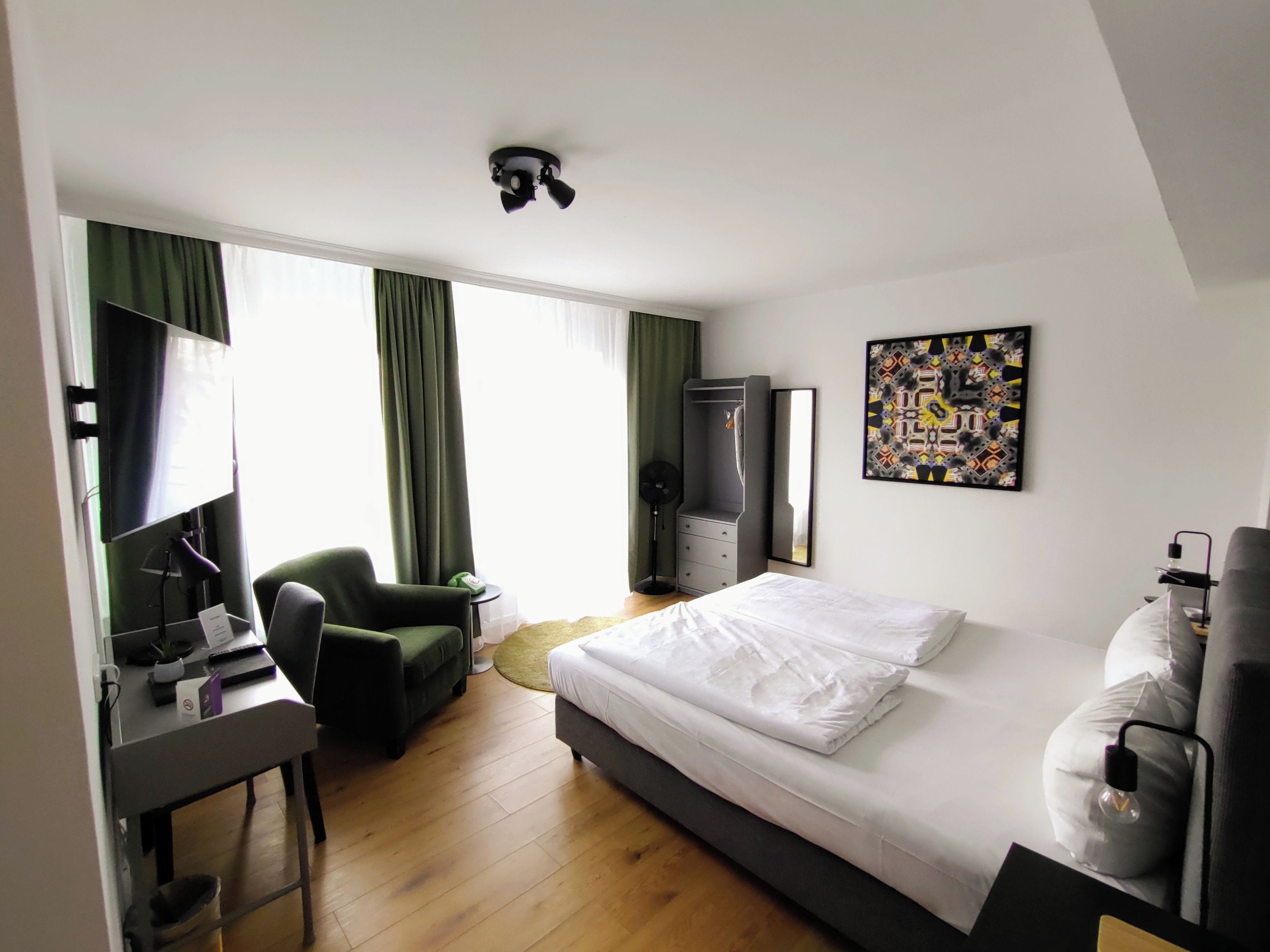 Habitación de hotel con cama, escritorio, silla, armario y un amplio ventanal con cortinas.