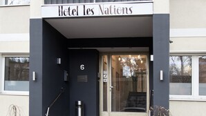 Entrada de un hotel con un letrero que dice "Hotel les Nations", un conjunto de escaleras con alfombra roja y dos grandes macetas con plantas.