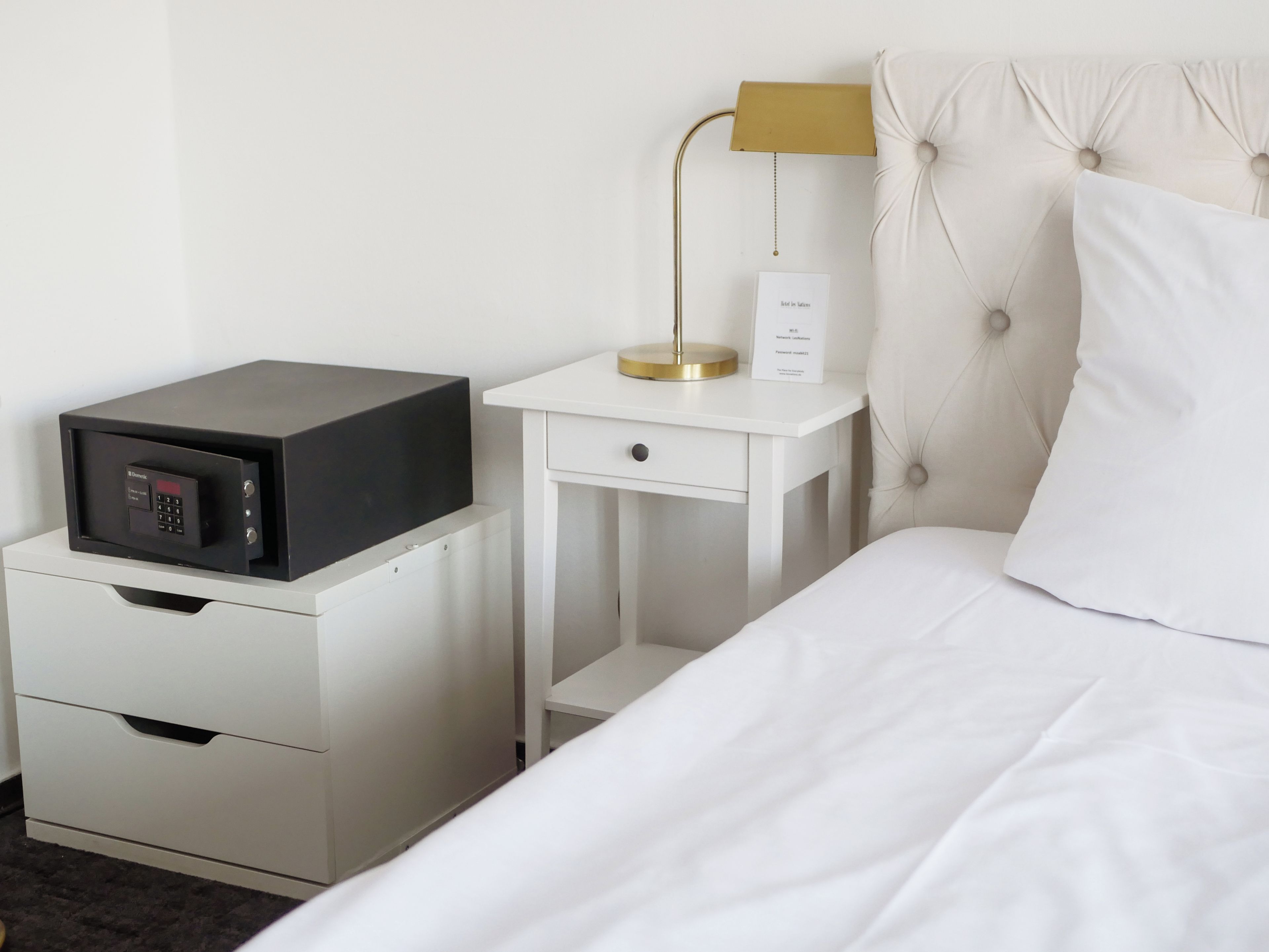 A hotel room with a bed, a bedside table with a lamp, a small desk with a computer, and a minibar.