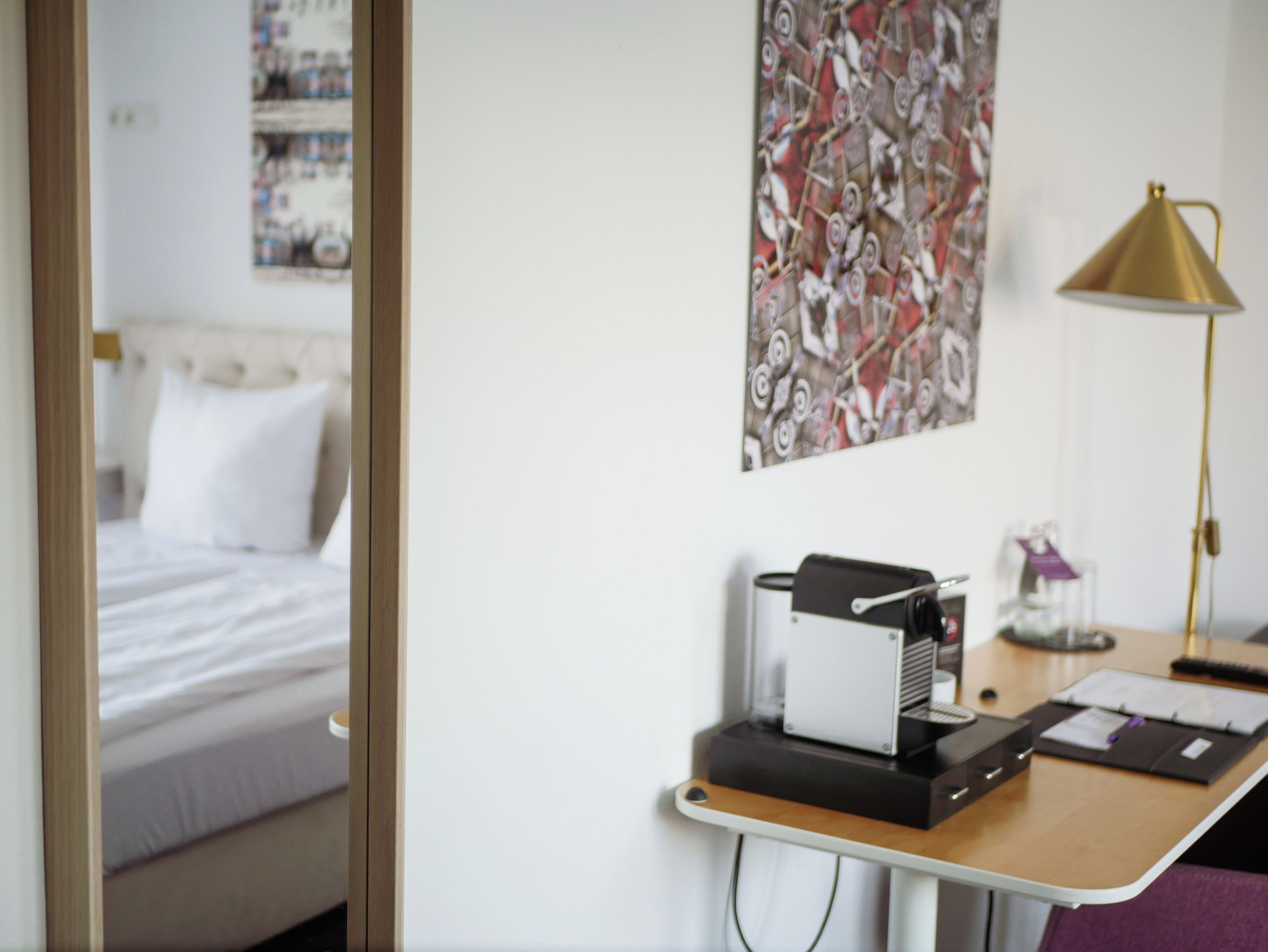 Ein Hotelzimmer mit einem Bett, einem Schreibtisch mit Drucker, einer Lampe und einem Wandbild mit abstrakten Motiven.