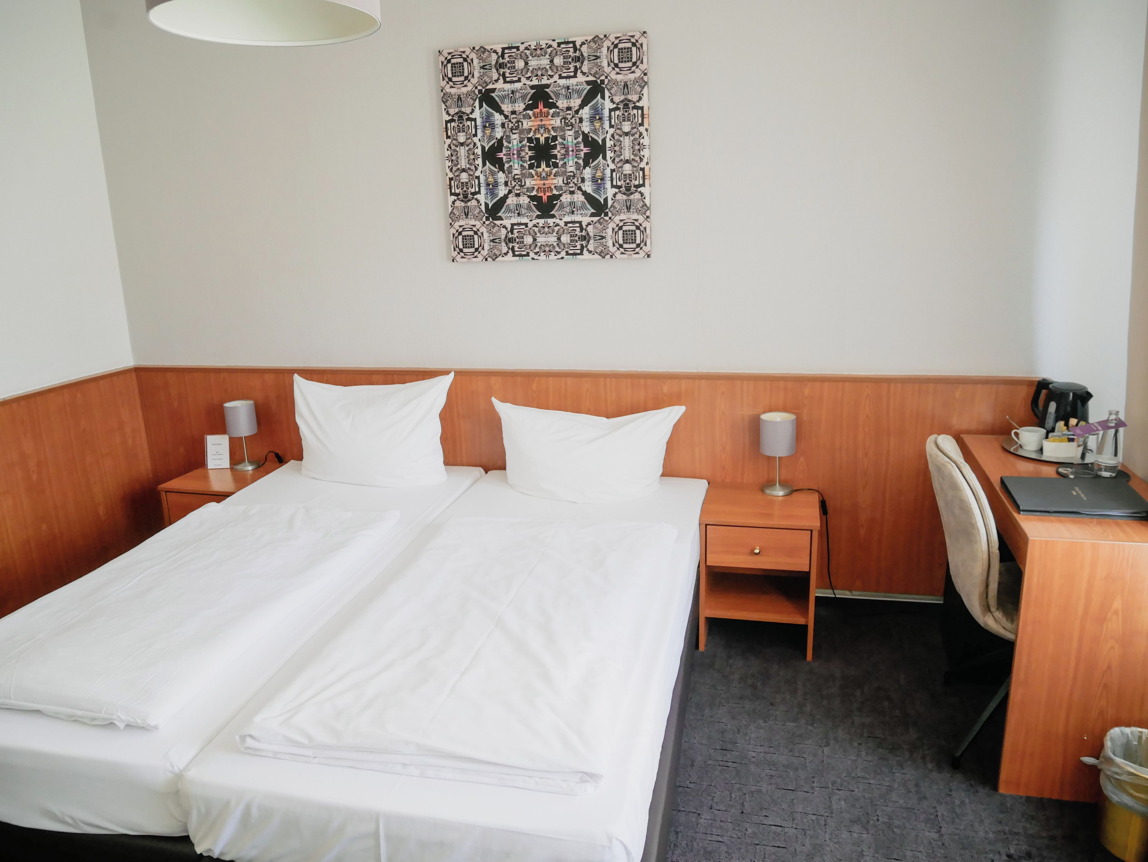 A hotel room with a double bed, two bedside tables, a desk, a chair, a lamp, and a tapestry on the wall.