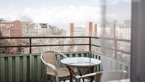 Un balcón con sillas de mimbre y una mesa redonda pequeña, con vistas a un paisaje urbano con edificios y árboles.