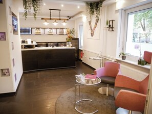 A reception area with a counter, seating, and a window with plants. - Hotel Les Nations (Berlin)