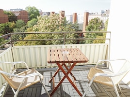 Un balcón con mesa de madera y dos sillas de mimbre, con vistas a un paisaje urbano con edificios y árboles.