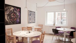 A dining area with round tables and chairs, a large abstract painting, and a window with a view of buildings. - Hotel Les Nations (Berlin)