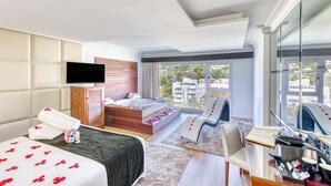 A modern hotel room with a large bed, a wooden headboard, a TV, a bathtub with rose petals, a desk with toiletries, and a city view. - Senator Marbella Hotel (Marbella)