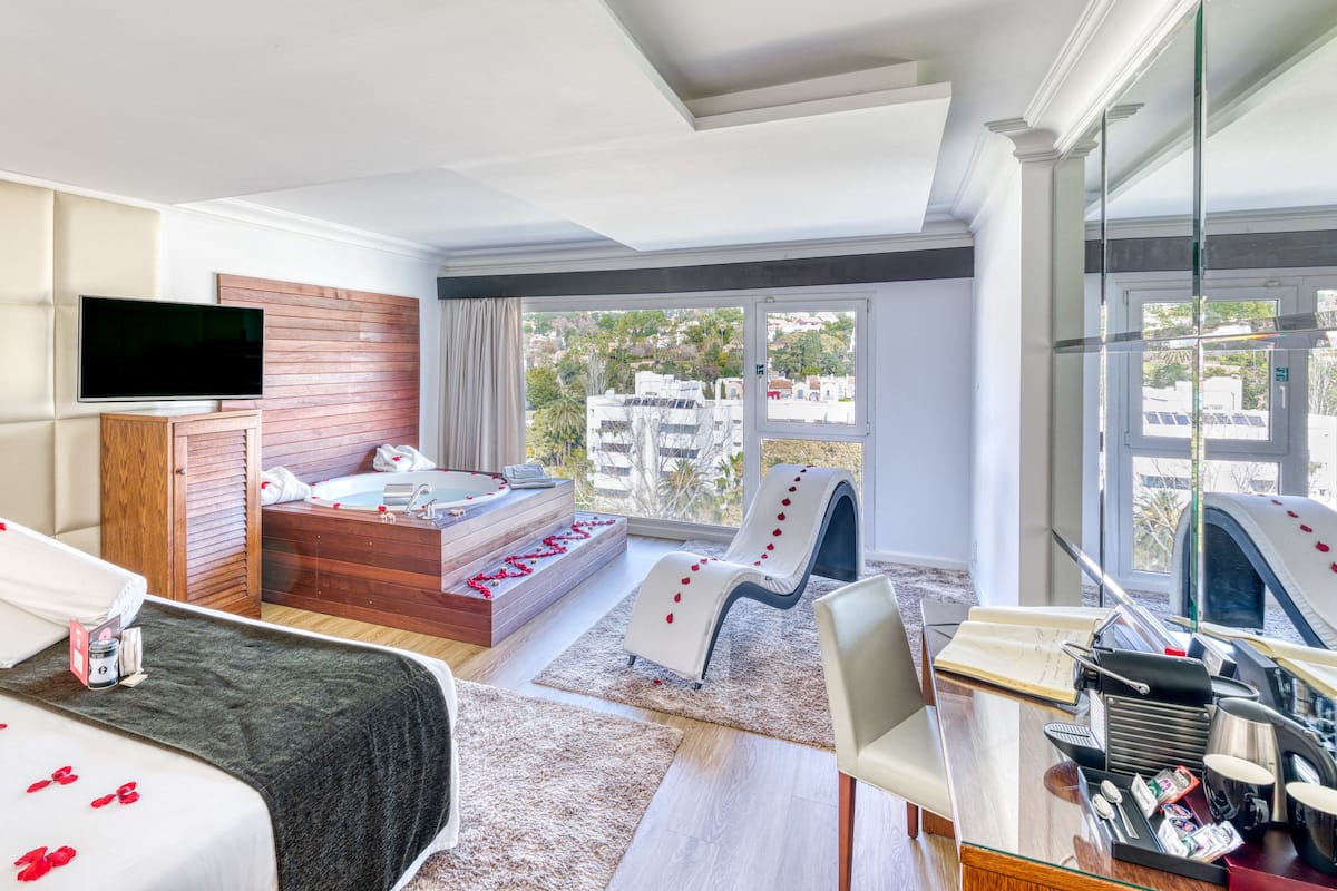 A modern hotel room with a large bed, a wooden headboard, a TV, a bathtub with rose petals, a desk with toiletries, and a city view.