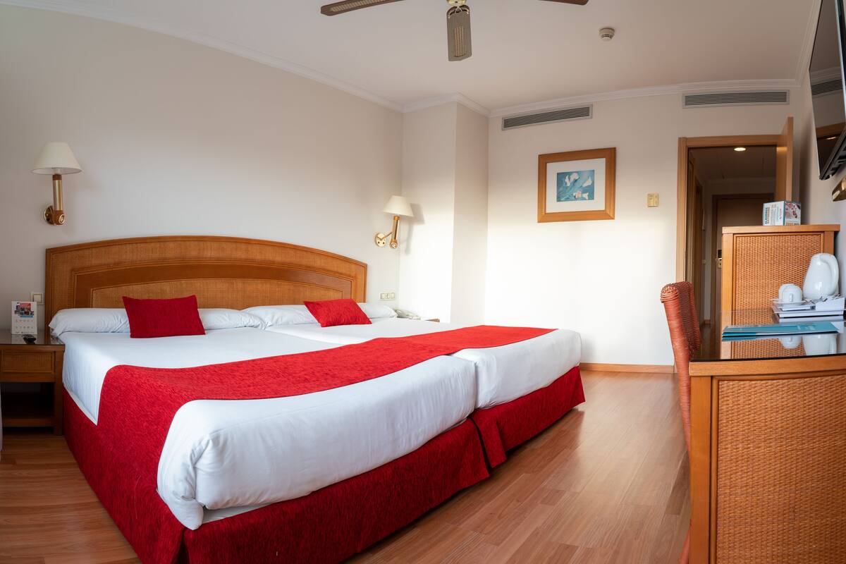 A hotel room with a large bed, wooden headboard, two bedside lamps, a desk with a chair, and a framed picture on the wall.