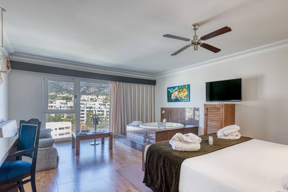 A hotel room with a bed, a desk, a chair, a TV, and a balcony with a view of buildings.