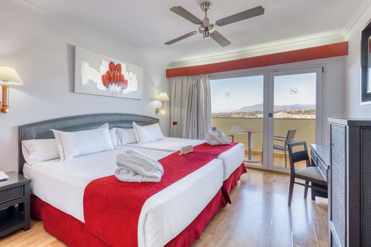 A hotel room with a large bed, a desk, a chair, a balcony with a view, and a ceiling fan.