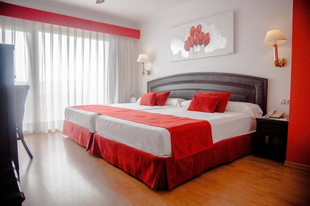 A double bed with red bedding, a bedside table with a lamp, and a painting on the wall.