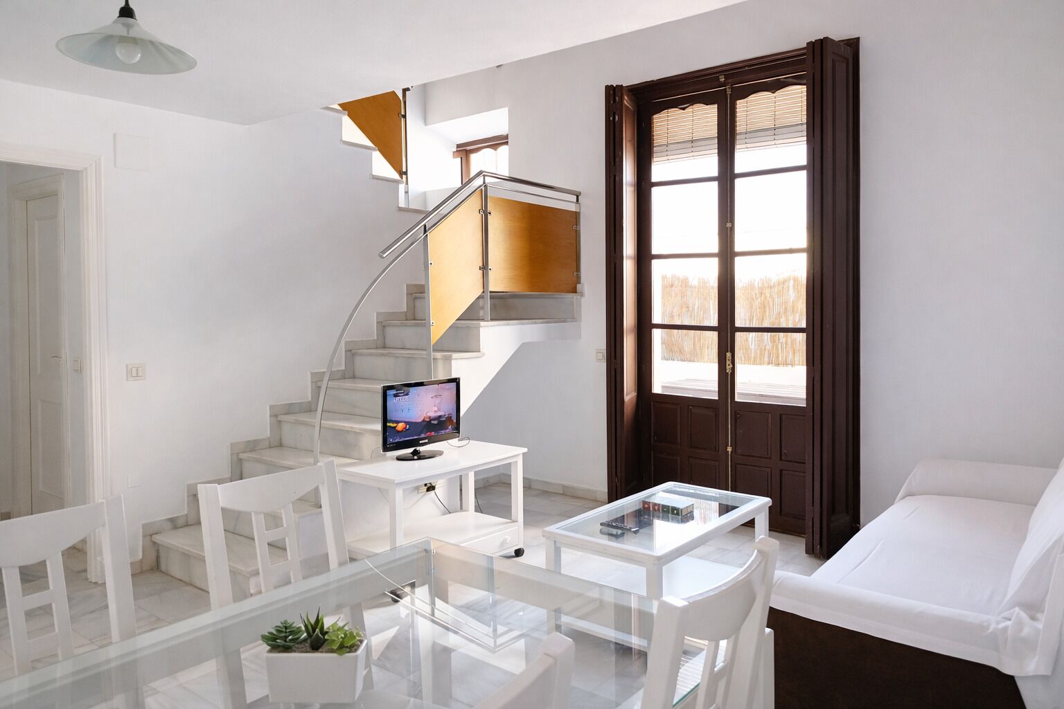 Una sala de estar moderna con un sofá blanco, una mesa de centro de vidrio, un televisor sobre un mueble blanco, una escalera con peldaños de madera y un ventanal con persianas.