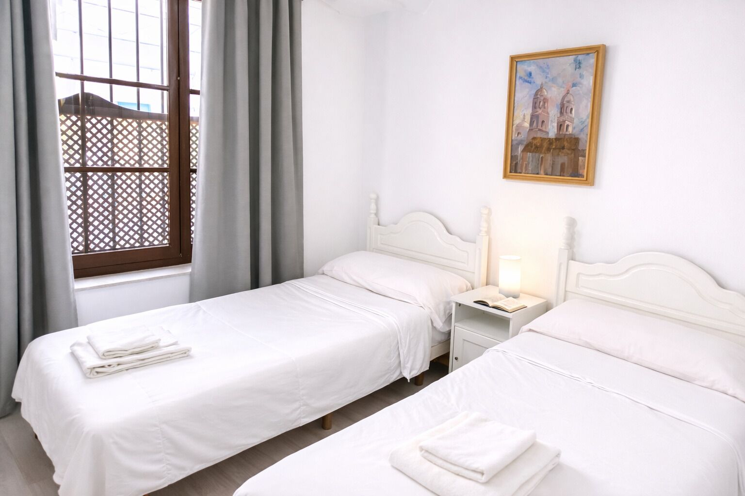 A hotel room with two beds, a window with curtains, a nightstand with a lamp, and a painting on the wall.