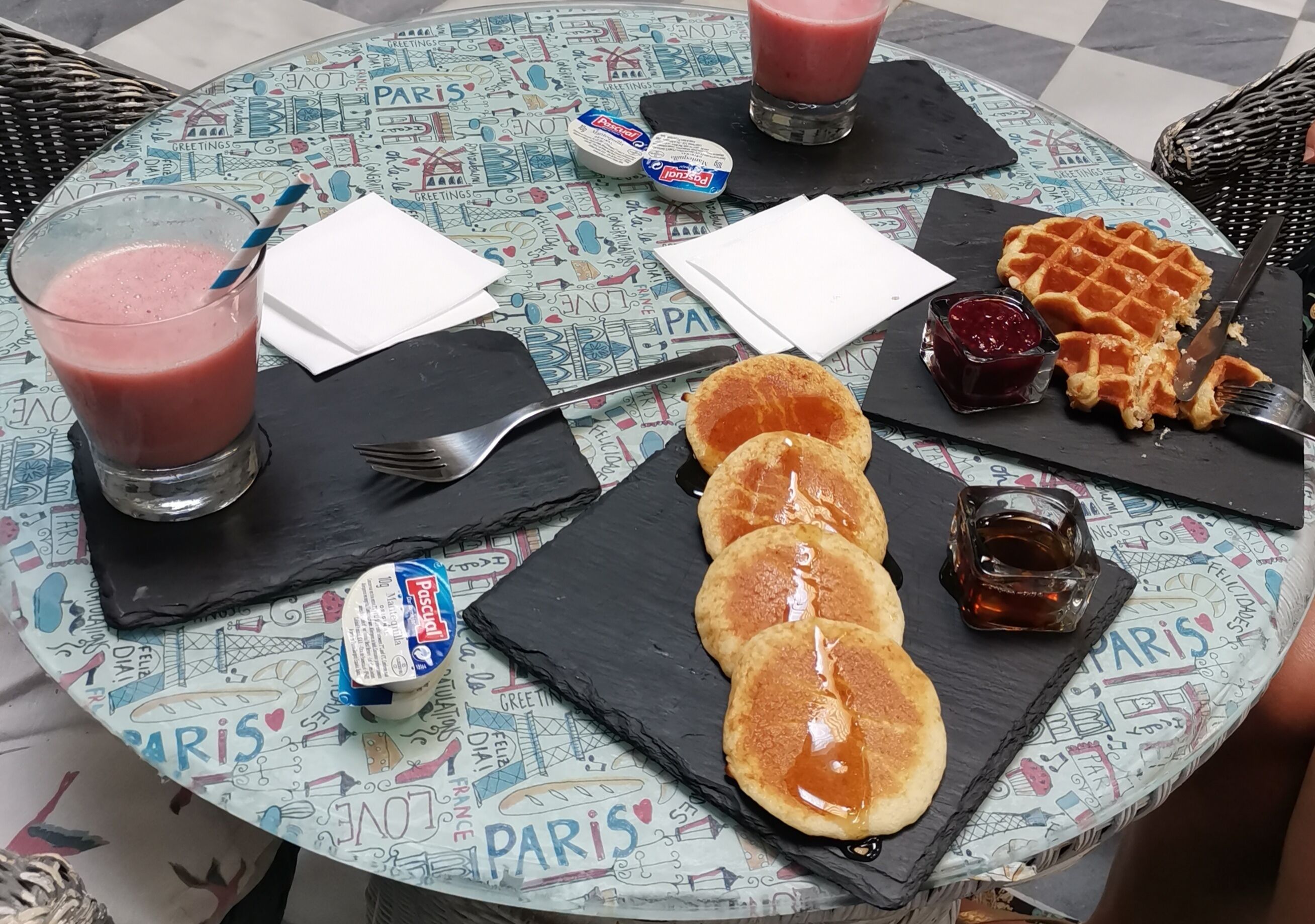 A round table with a Paris-themed tablecloth, a glass of pink drink, a waffle, pancakes, and condiments.