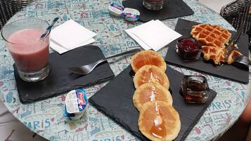 A round table with a Paris-themed tablecloth, a glass of pink drink, a waffle, pancakes, and condiments.