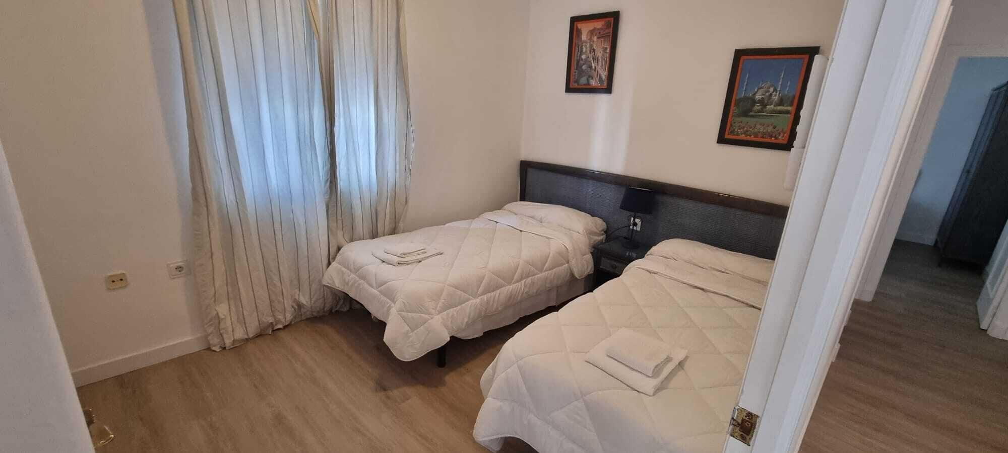 A room with two beds, a window with curtains, and two framed pictures on the wall.