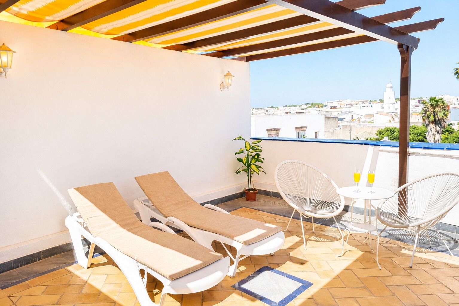 A terrace with two sun loungers, a potted plant, two wicker chairs, and a table with two glasses of orange juice.