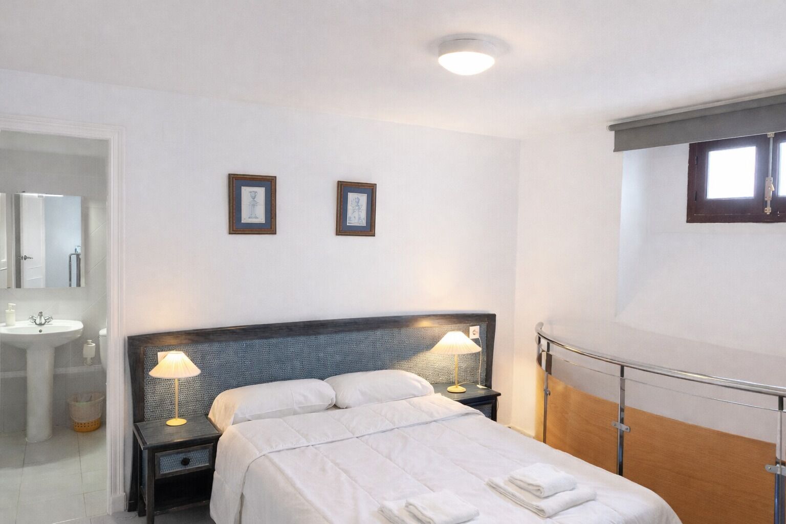 Habitación de hotel con cama, lámparas de noche, mesita, espejo, lavamanos y cuadros en la pared.