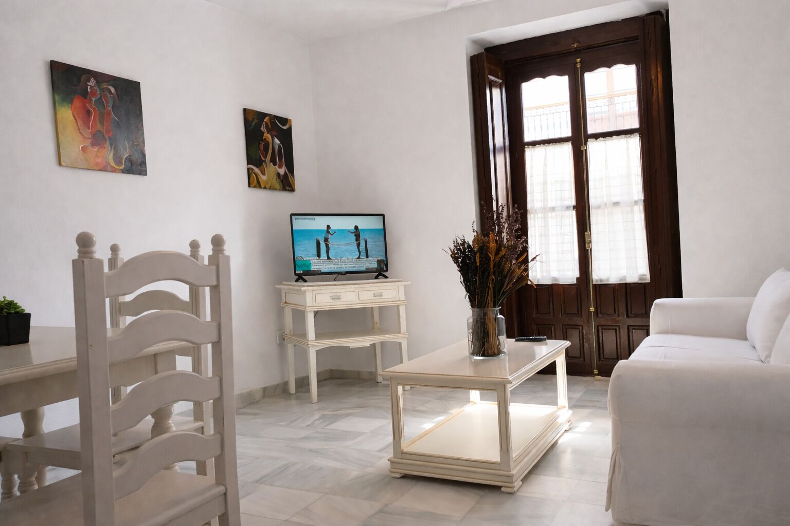 A living room with a TV, white furniture, a vase with dried plants, and a door with a window.