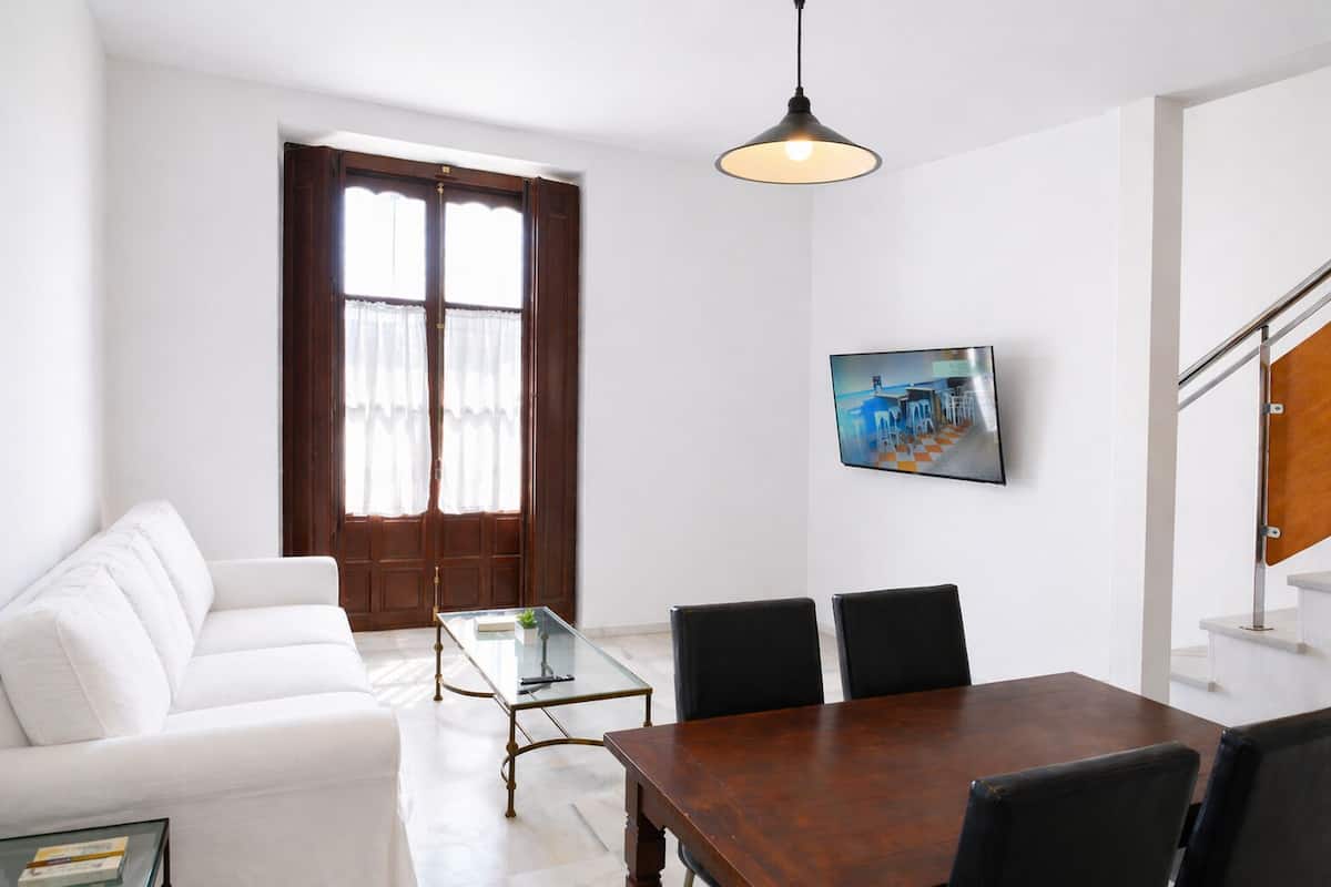 Een moderne woonkamer met een witte bank, een glazen salontafel, een houten eettafel met stoelen, een groot raam met gordijnen en een flatscreen televisie aan de muur.