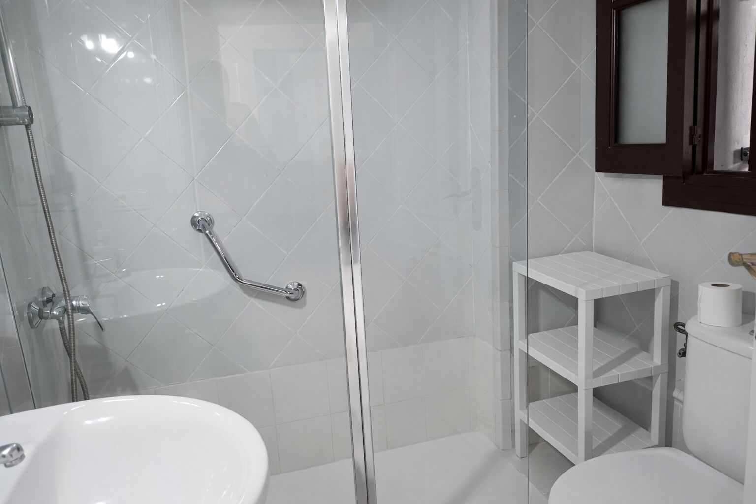 Un baño con ducha envuelta en vidrio, un lavamanos blanco, un inodoro y un pequeño estante blanco con un rollo de papel higiénico.