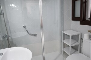 Un baño con ducha envuelta en vidrio, un lavamanos blanco, un inodoro y un pequeño estante blanco con un rollo de papel higiénico.