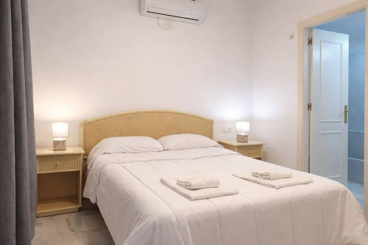 Een keurig opgemaakt bed met witte lakens en twee opgevouwen handdoeken, een bedlamp op elk nachtkastje, en een airconditioner boven het bed.