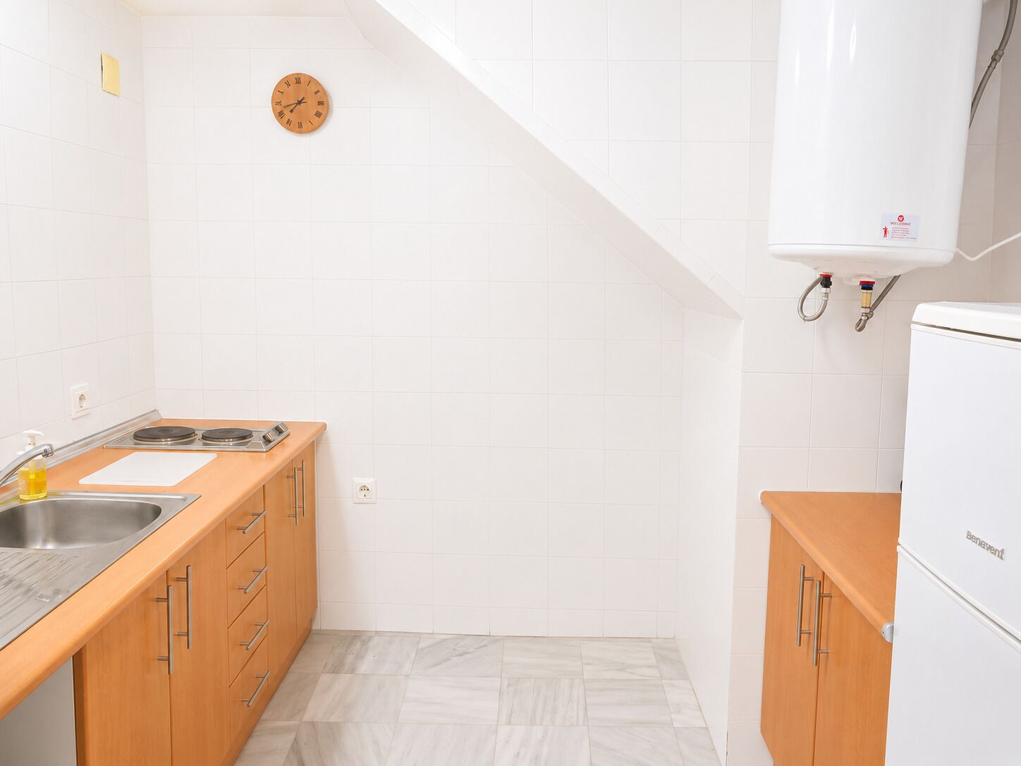 Une petite cuisine équipée d’un évier, d’une cuisinière, de placards en bois et d’une horloge au mur.