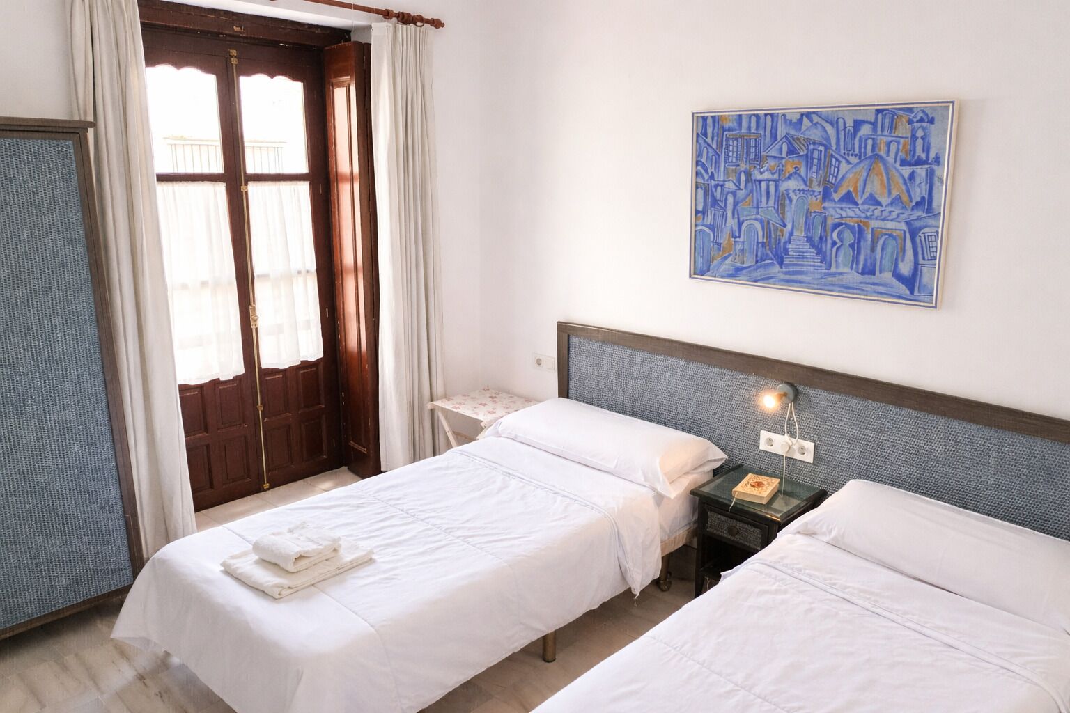 A hotel room with two single beds, a wooden door, a window with curtains, a bedside table with a lamp, and a painting on the wall.