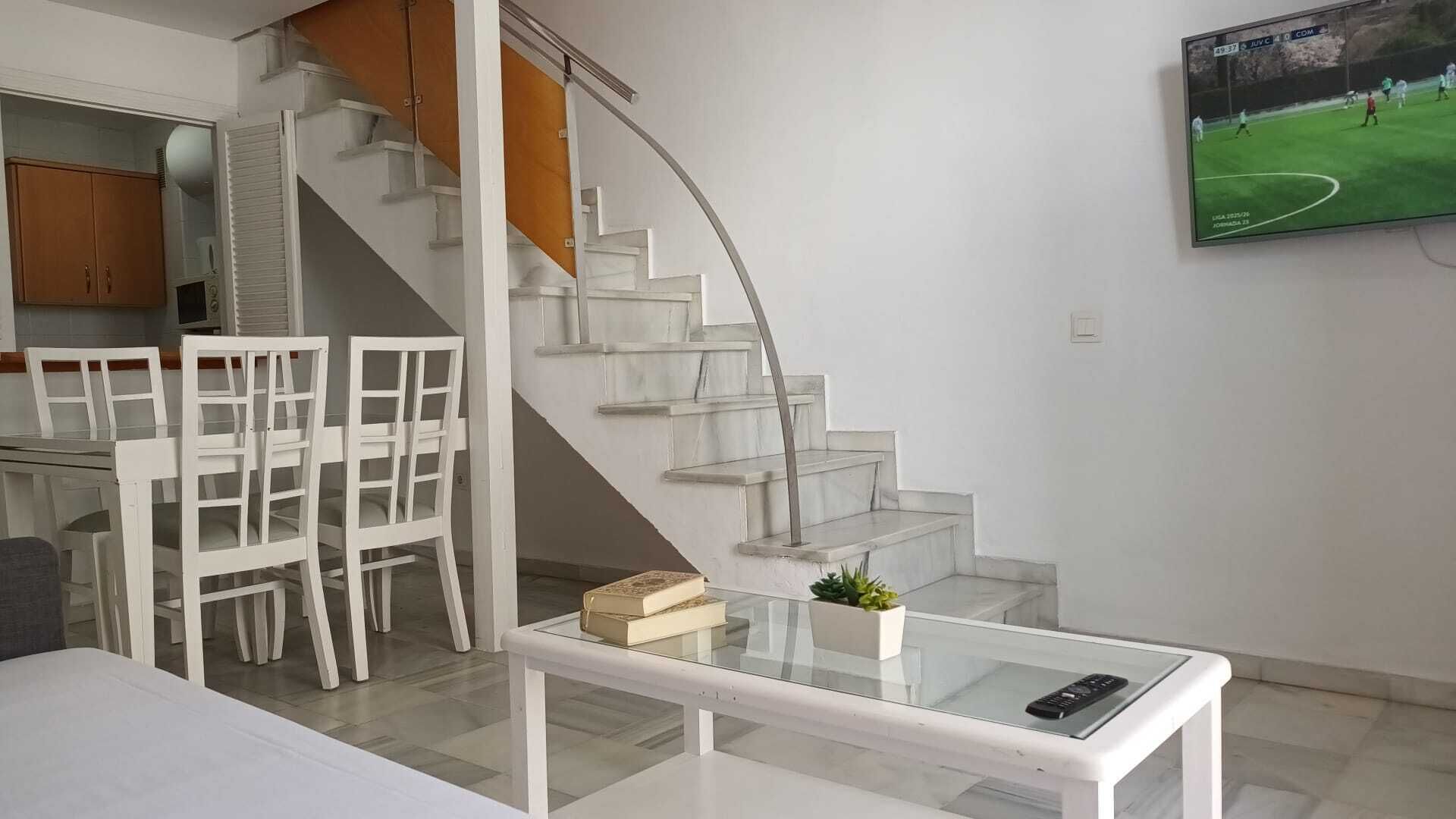 Un salon moderne avec une table basse en verre, des meubles blancs, un escalier et un téléviseur fixé au mur.