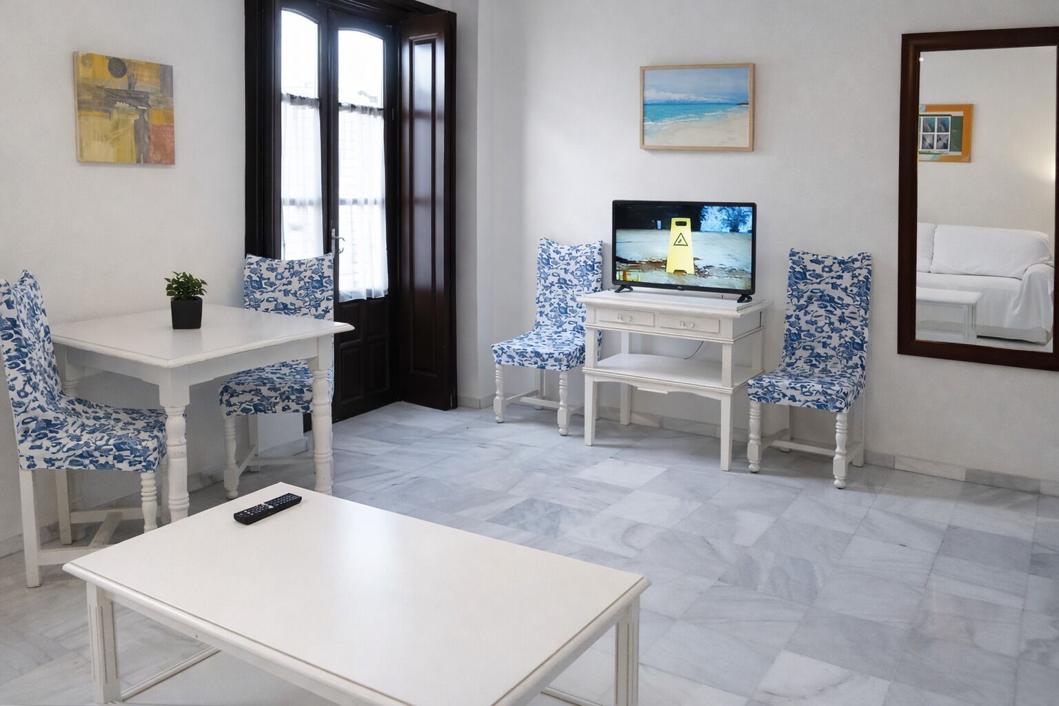 Une pièce avec une table blanche, deux chaises recouvertes d’un tissu à motifs floraux bleus, un téléviseur posé sur un meuble blanc, une petite plante en pot et un tableau accroché au mur.