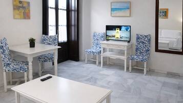 Une pièce avec une table blanche, deux chaises recouvertes d’un tissu à motifs floraux bleus, un téléviseur posé sur un meuble blanc, une petite plante en pot et un tableau accroché au mur.