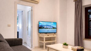 Una sala de estar con un televisor sobre un mueble bajo, un sofá, una mesita con una planta y libros, y una puerta que da a otra habitación.