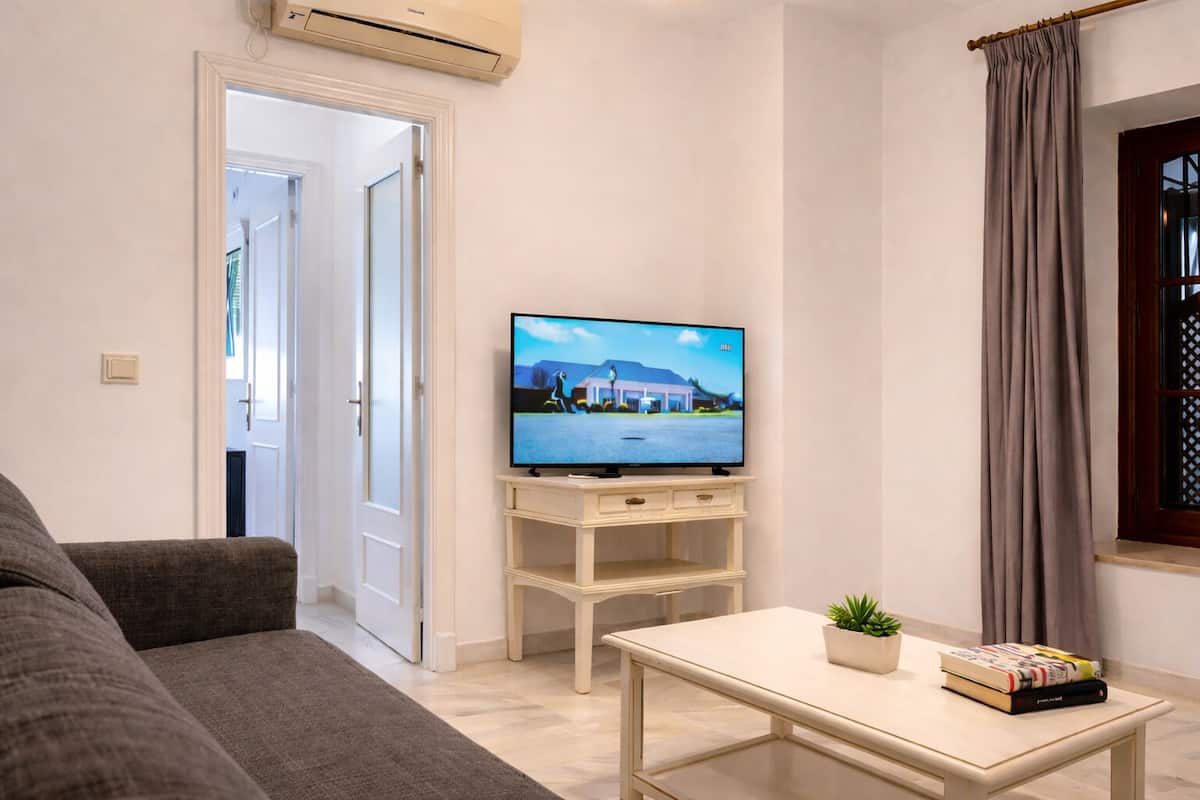 Een woonkamer met een televisie op een standaard, een bank, een kleine tafel met een plant en boeken, en een deur die leidt naar een andere kamer.