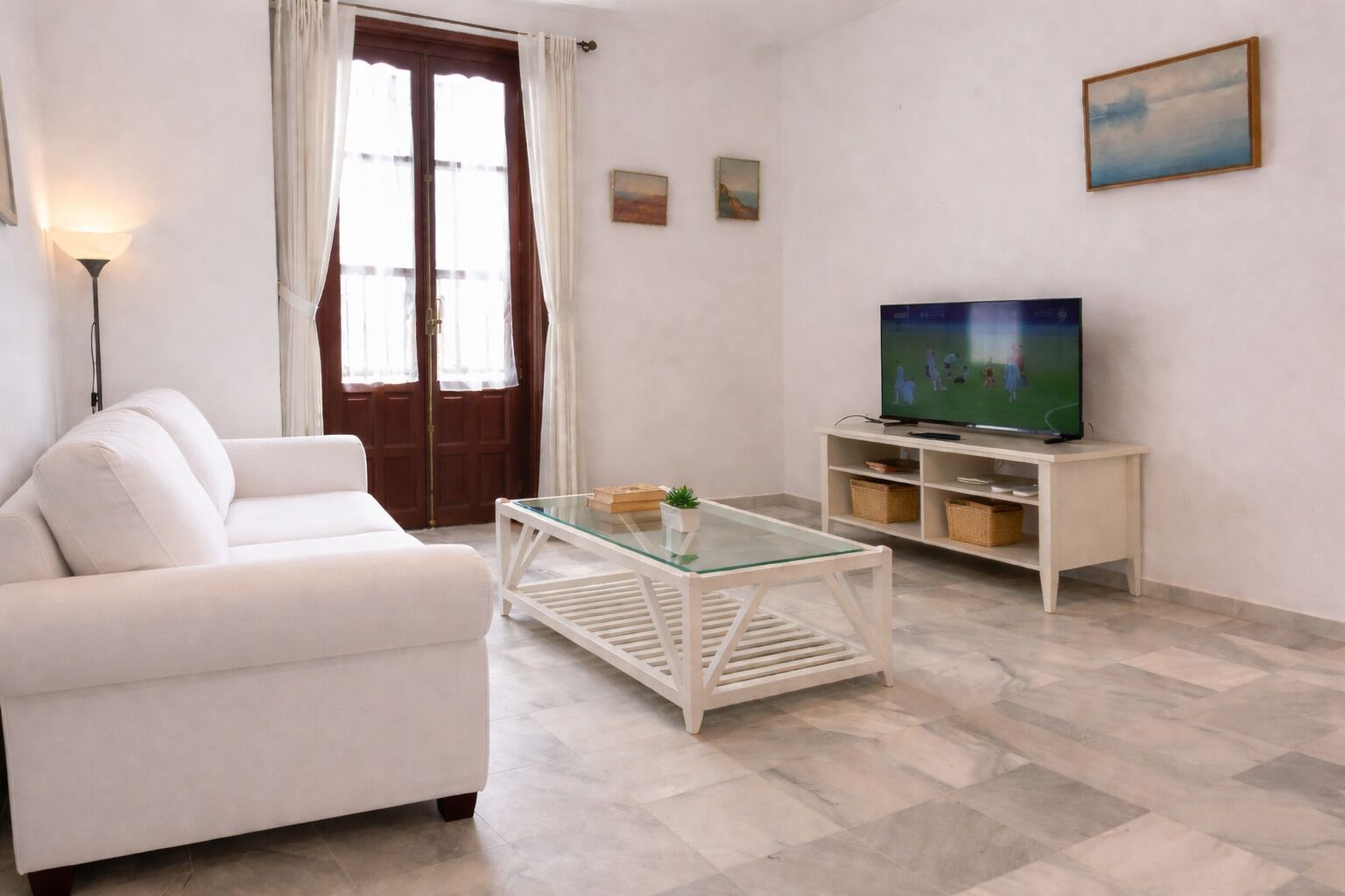 A living room with a white sofa, a glass coffee table, a TV on a wooden stand, and a door with curtains.