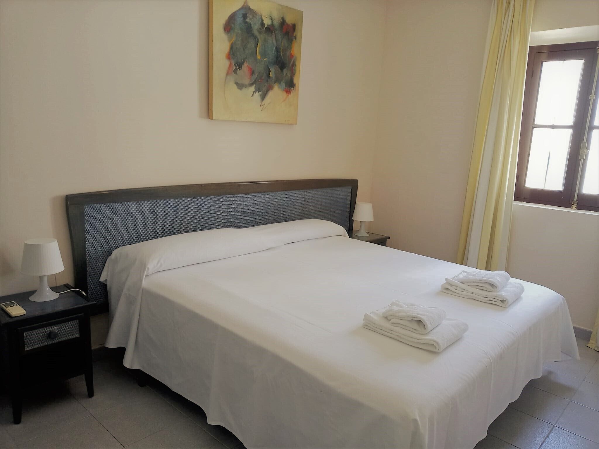 Una cama bien hecha con ropa y toallas blancas, una lámpara de noche, una mesita con lámpara y un cuadro en la pared.