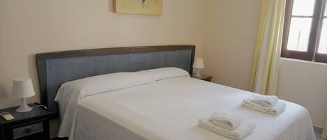 Una cama bien hecha con ropa y toallas blancas, una lámpara de noche, una mesita con lámpara y un cuadro en la pared.