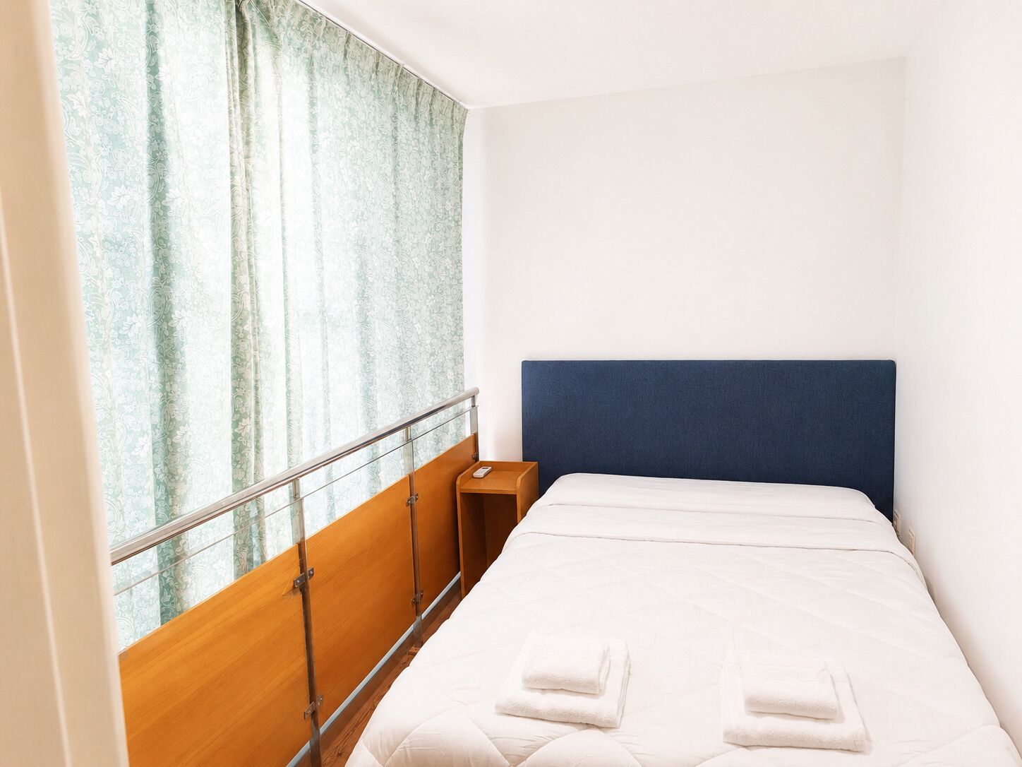 A hotel room with a bed, a nightstand, a window with curtains, and a wooden headboard.