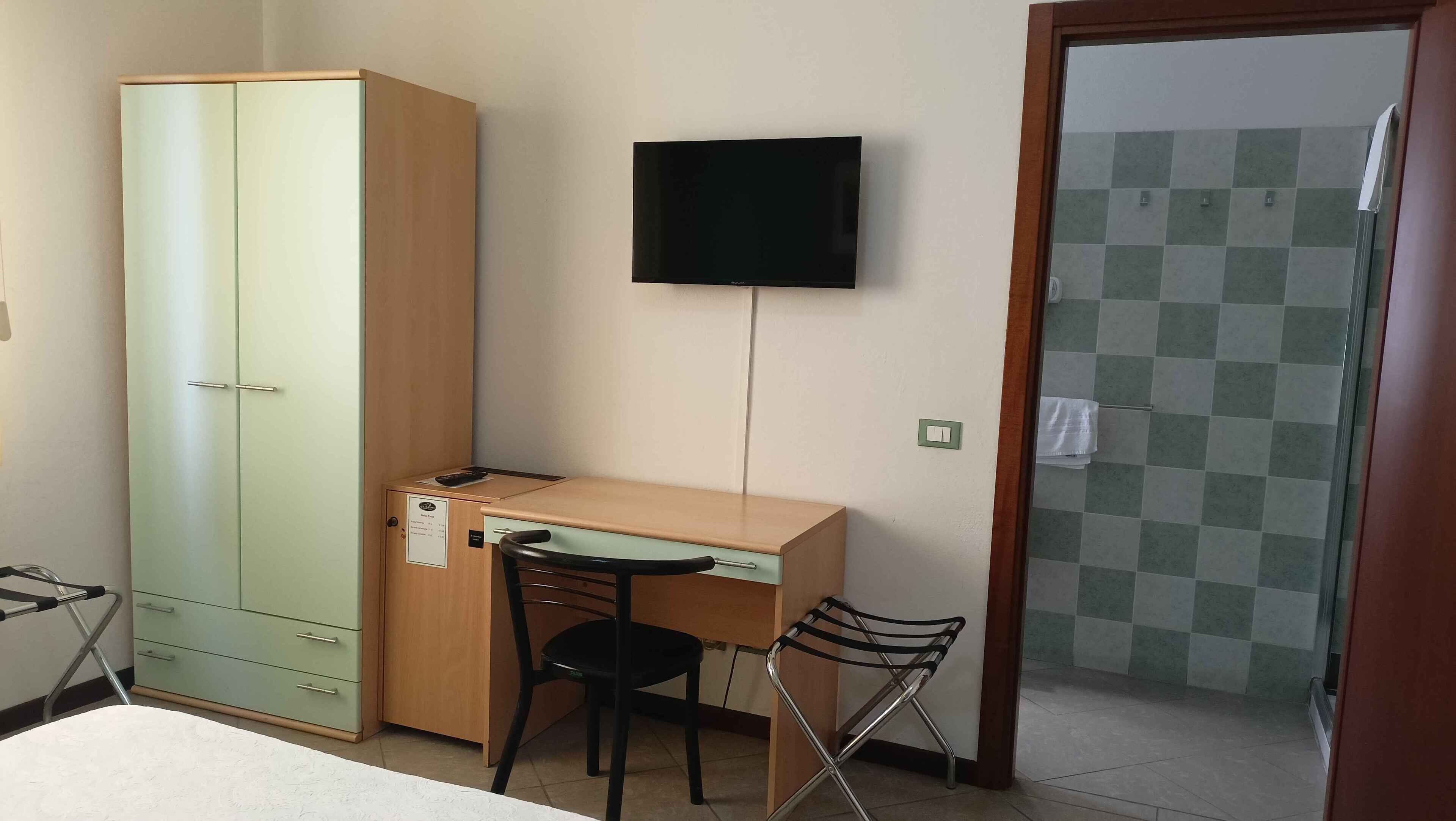 Une petite pièce comprenant un bureau, une chaise et une armoire. Un téléviseur est fixé au mur, et une porte donne accès à une salle de bain dont le sol est carrelé.