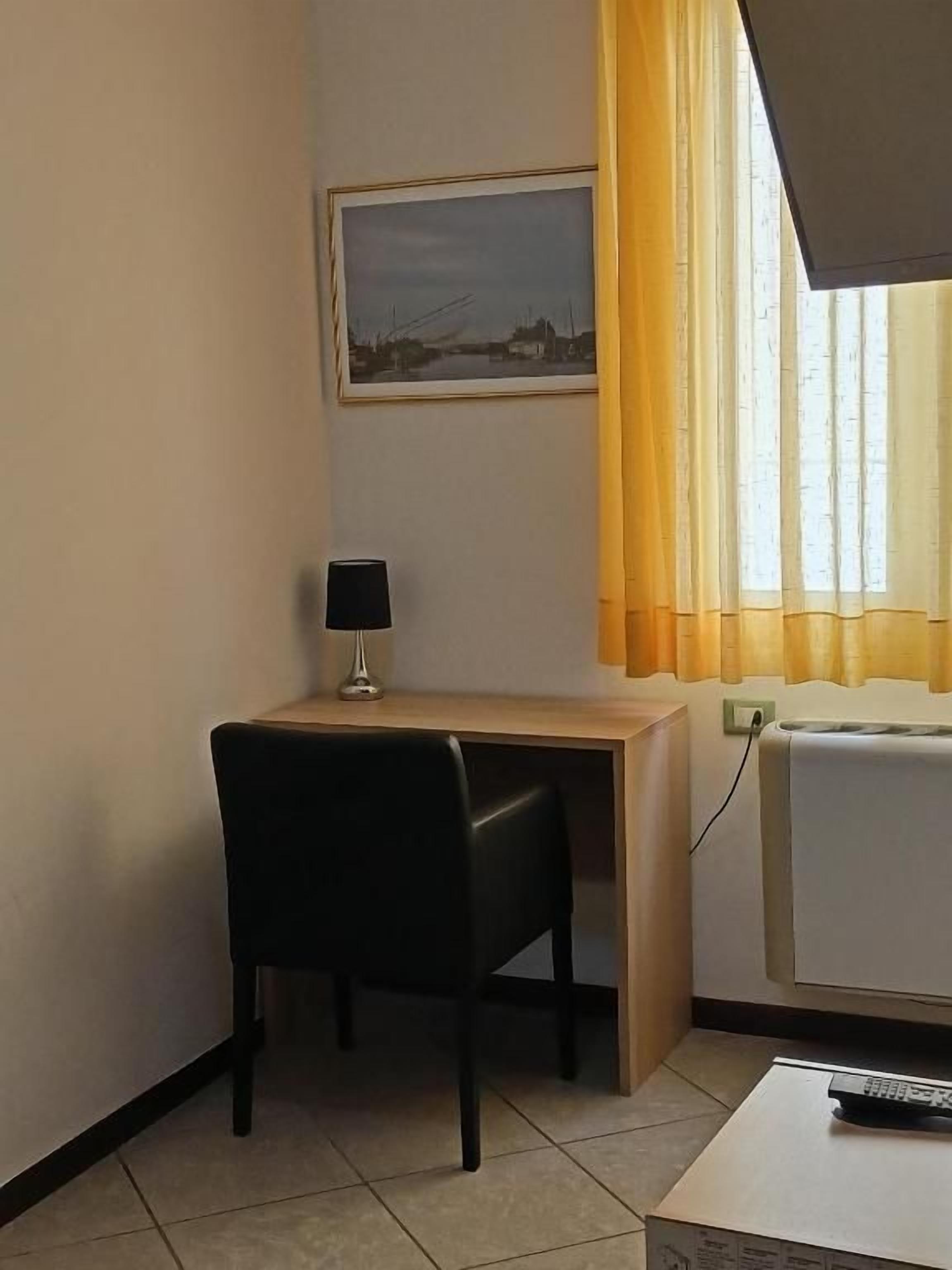 Ein kleines Zimmer mit Schreibtisch, Stuhl, Lampe, einem Fenster mit Vorhängen und einem Fernseher.