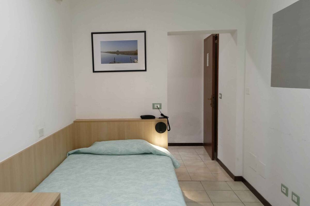 A small, simple room with a bed, a desk, and a chair. There is a framed picture on the wall and a door leading to another room.